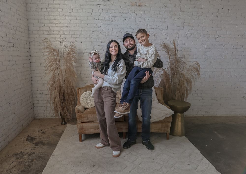 Family of four posing together in a cozy studio with white brick walls, pampas grass decor, and warm neutral tones. Minot, ND photographer