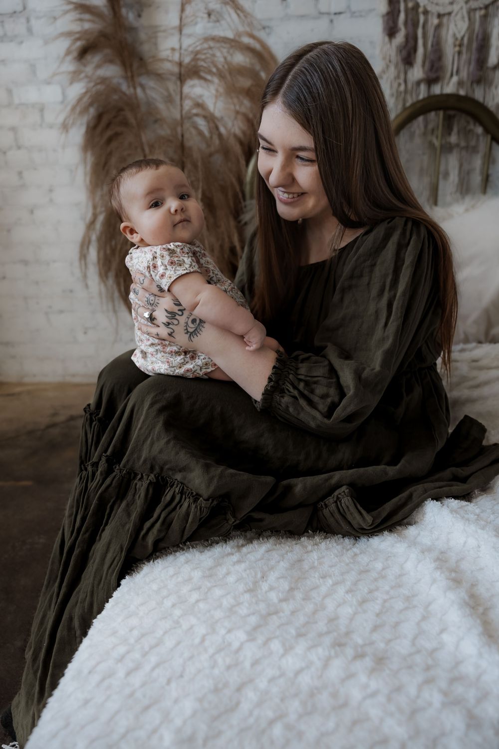 Smiling young mother in dark green dress holding her baby infant while sitting on a white fluffy surface. Minot, ND photographer