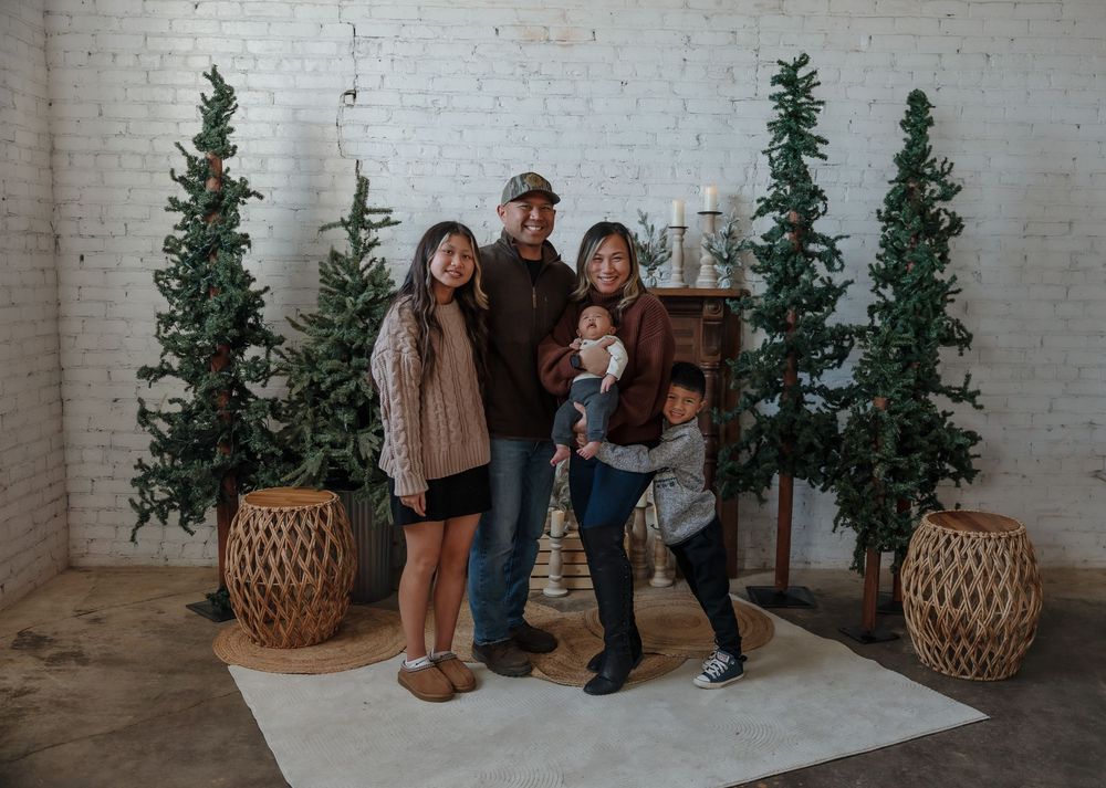 Family of five posing together in a festive studio with tall Christmas trees and white brick backdrop. Minot, ND photographer