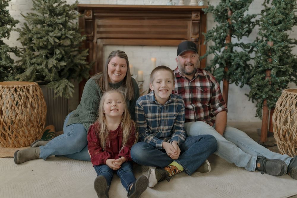 Family of four smiling together in front of Christmas trees and fireplace for holiday portrait photo. Minot, ND photographer