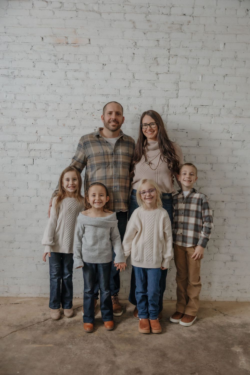 Family of six posing together in front of a white brick wall, wearing coordinated neutral and plaid outfits. Minot, ND photographer