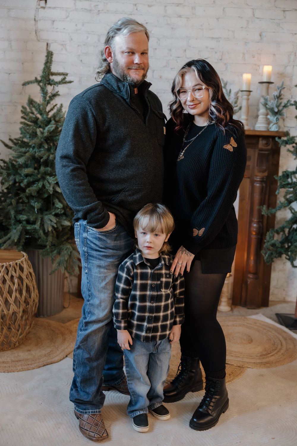 Family of three posing for a holiday portrait with Christmas trees and a fireplace in a cozy studio setting. Minot, ND photographer