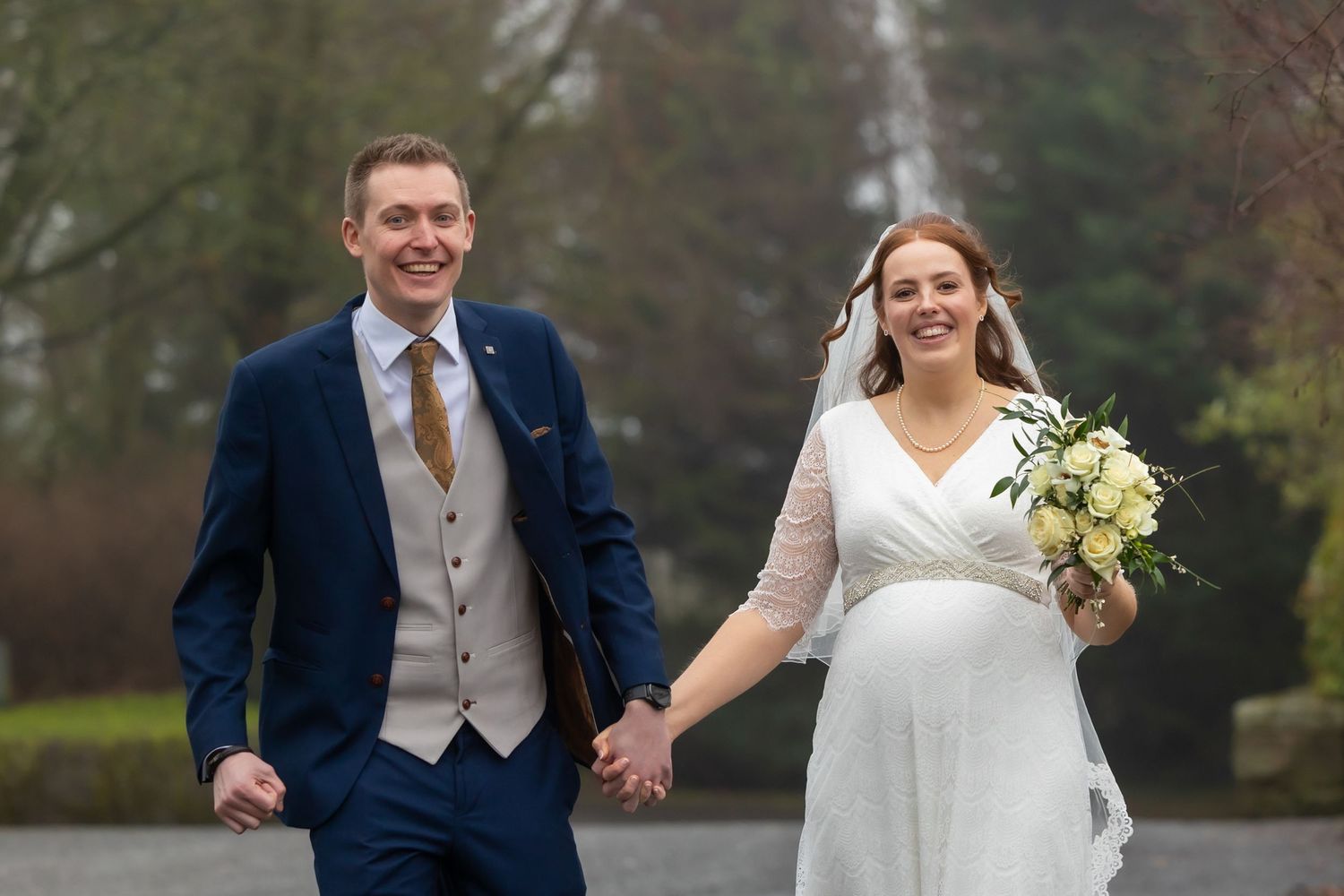 Smiling bride and groom holding hands walking outdoors, bride in white gown holding bouquet, groom in navy suit. Casual Captures Wedding
