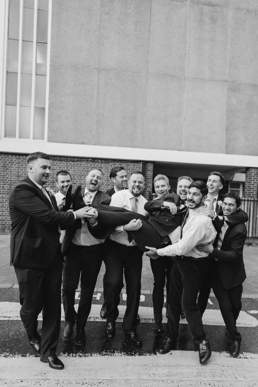 Black and white photo of groomsmen lifting and carrying the groom outdoors, all dressed in suits, laughing joyfully.