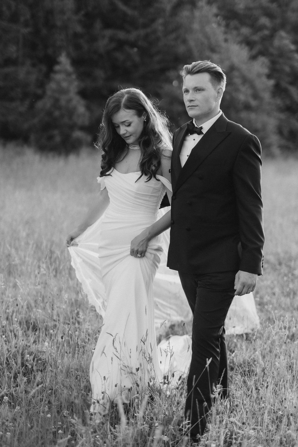 Black and white wedding photo of bride in flowing gown and groom in tuxedo walking through a meadow.