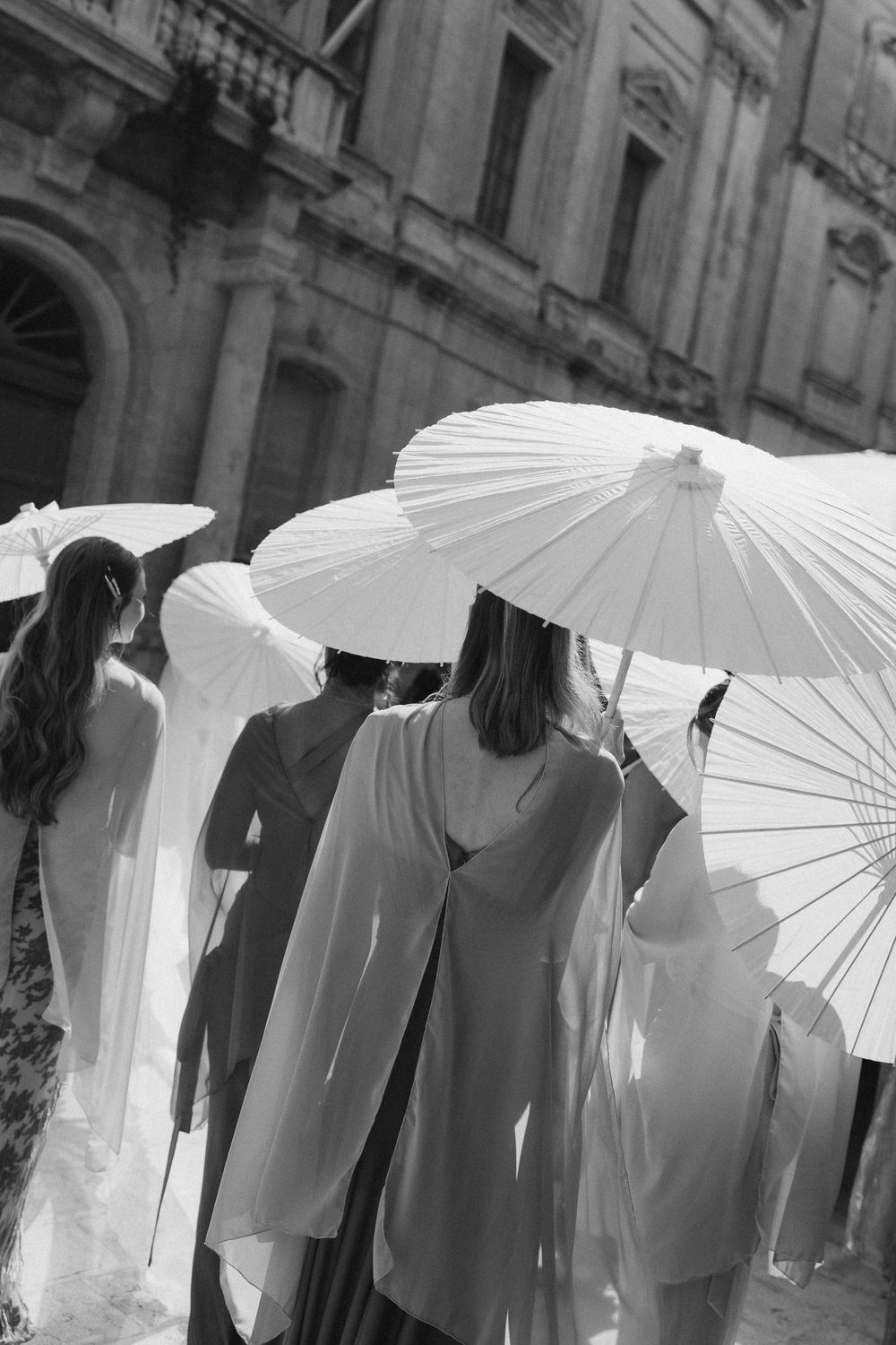 Black and white photo of women in elegant dresses holding white parasols on a sunlit European street.