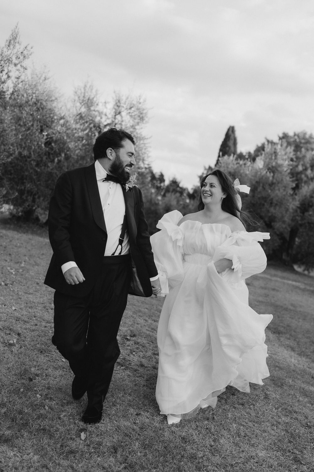 Black and white photo of a smiling bride and groom holding hands, walking outdoors on their wedding day in Tuscany.