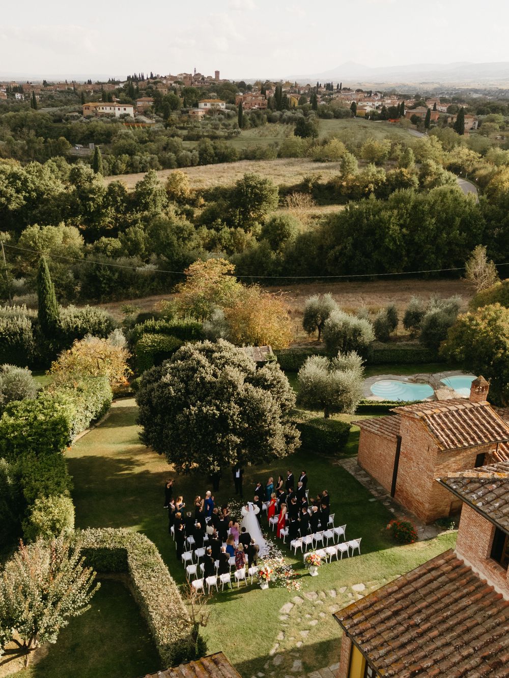Aerial view of an outdoor wedding ceremony in a lush Tuscan garden with rolling hills and a medieval town backdrop.