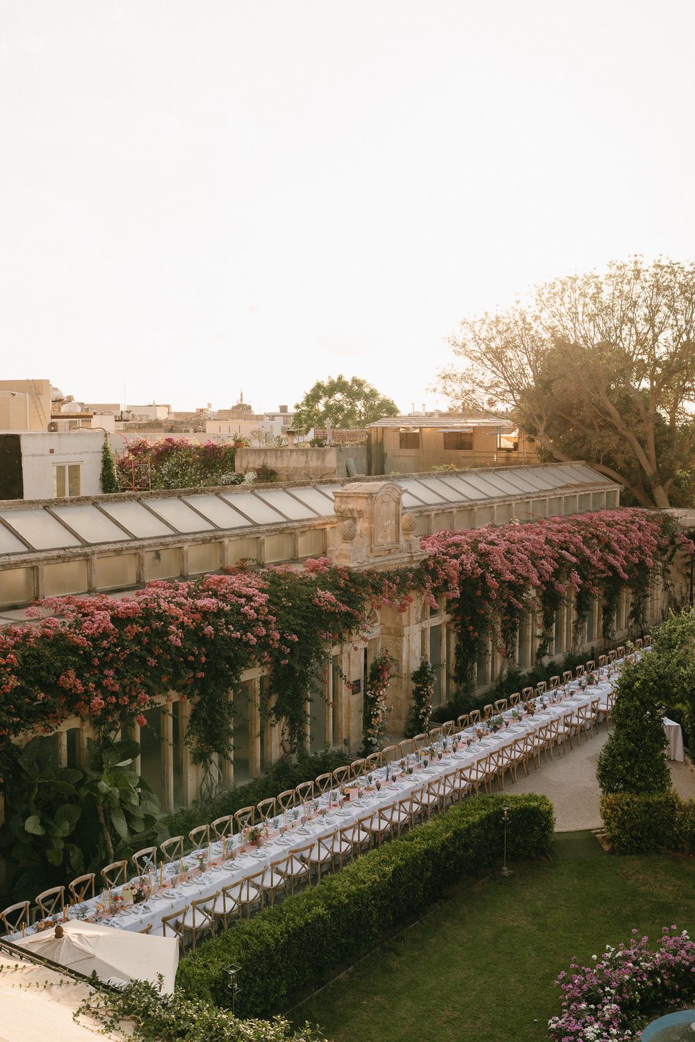 Elegant outdoor wedding reception with long dining tables in a garden beside a historic greenhouse adorned with pink bougainvillea.