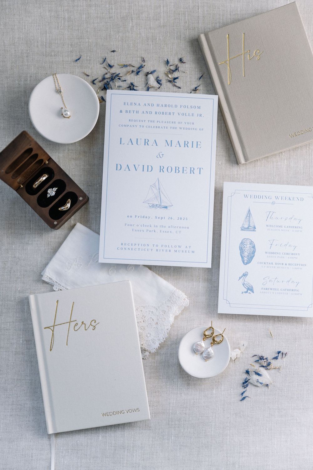 Elegant nautical-themed wedding invitation suite with blue accents, vow books, rings, and floral details on gray linen.