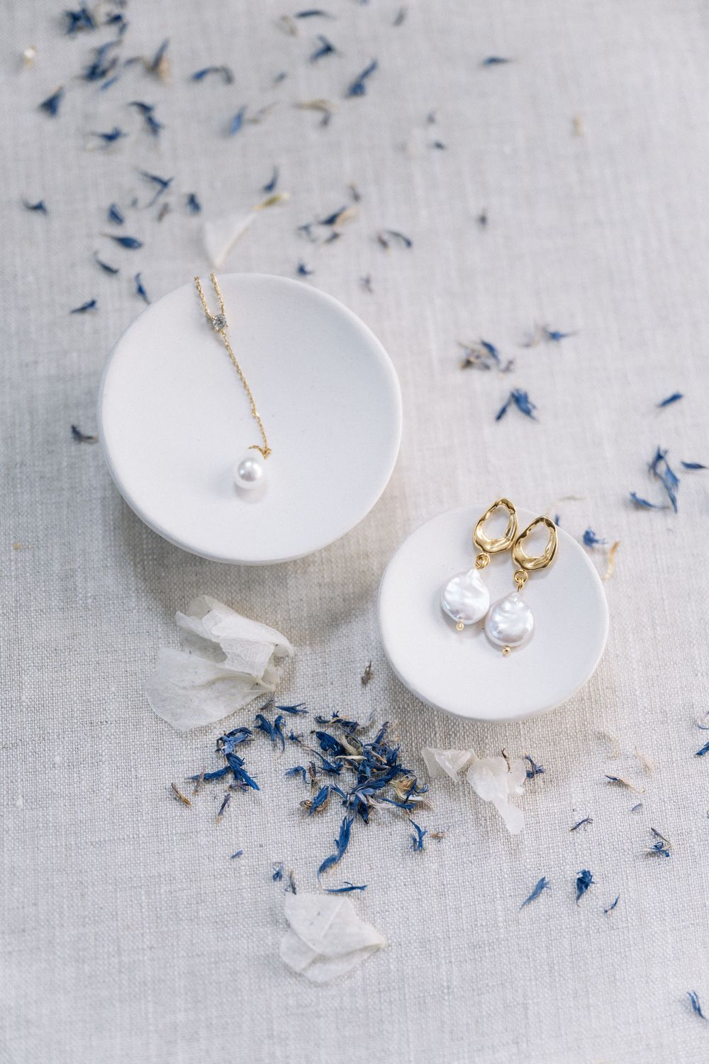 Pearl necklace and baroque pearl gold earrings arranged on white ceramic dishes with scattered blue petals.