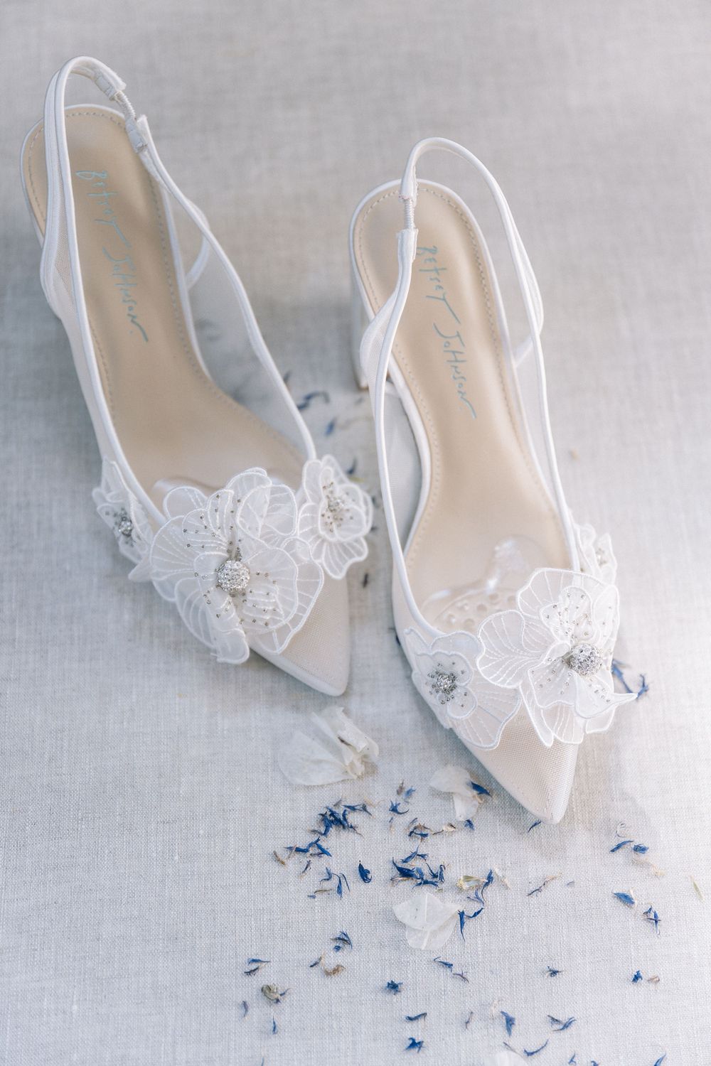 White lace slingback bridal heels with floral embellishments and crystal details on a soft gray background.