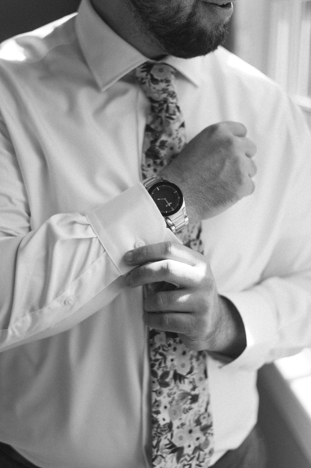 Groom in white dress shirt and floral tie adjusting his watch cufflink in black and white photo.
