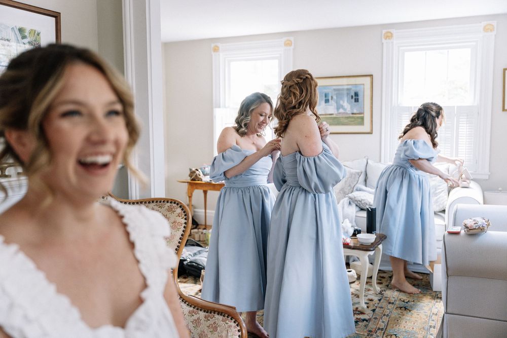 Bride laughing in white lace gown while bridesmaids in blue dresses help each other get ready indoors.