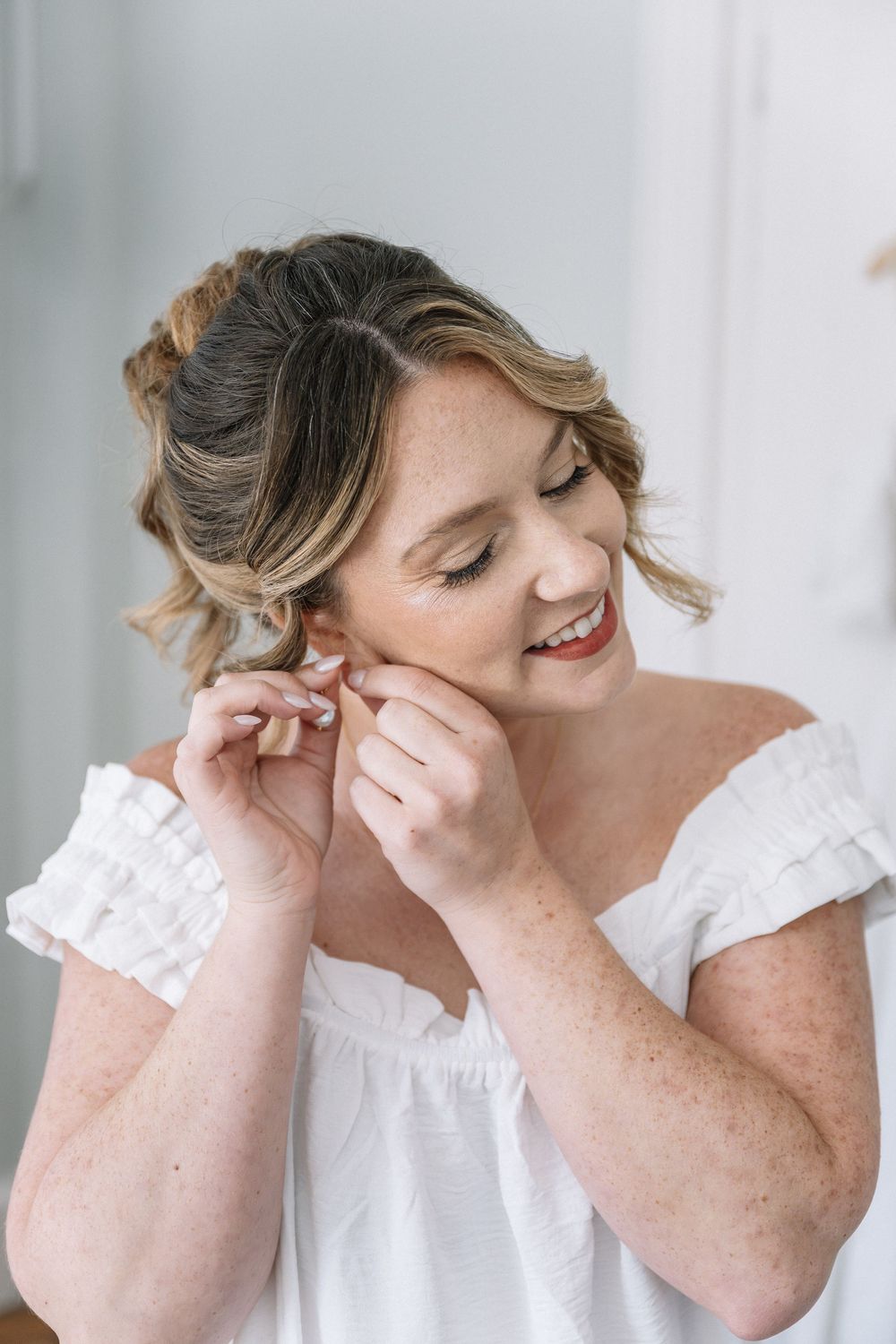 Smiling bride in white off-shoulder dress putting on earrings during wedding preparations.