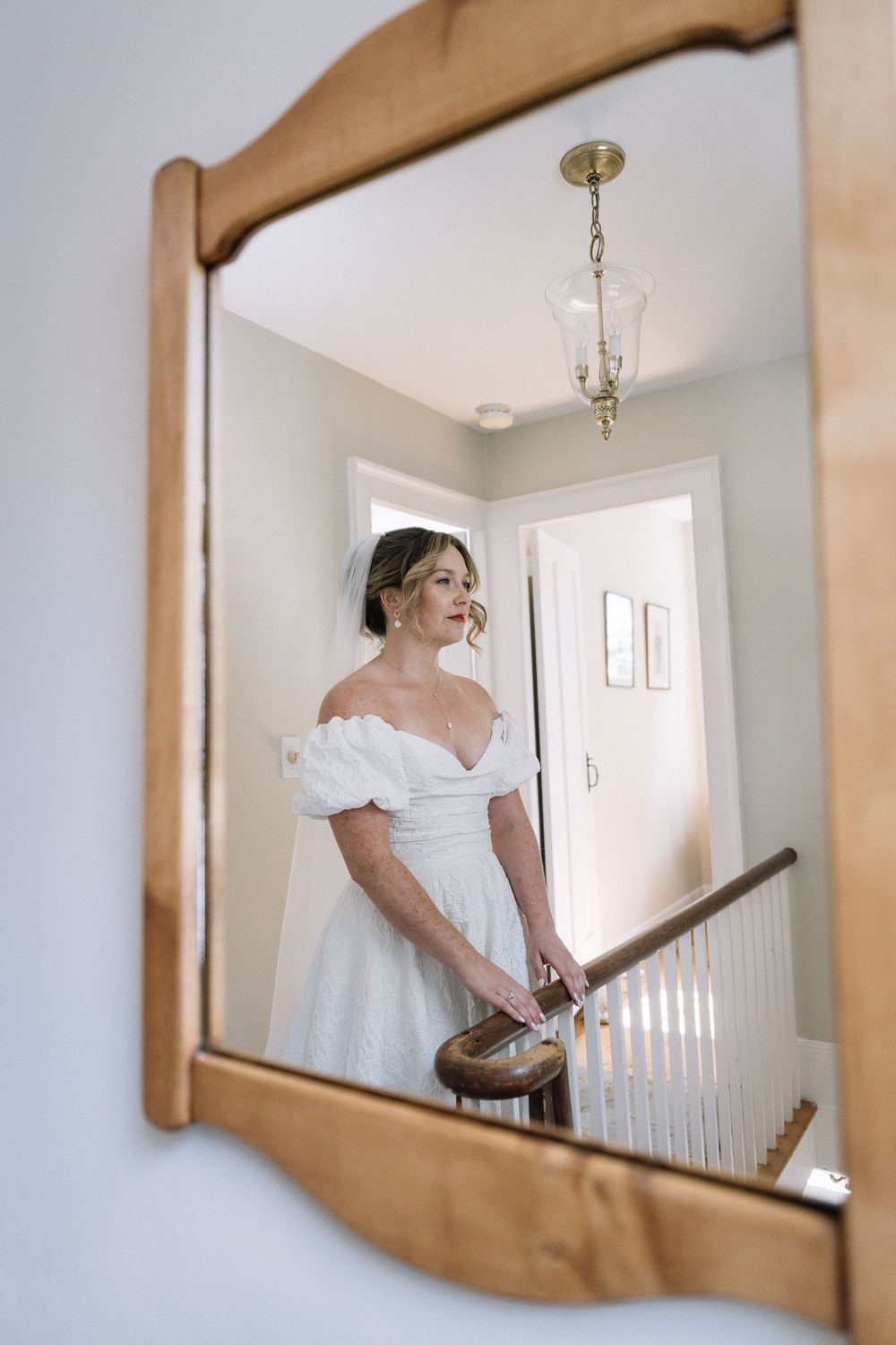 Bride in white off-shoulder wedding dress reflected in wooden mirror at top of staircase.