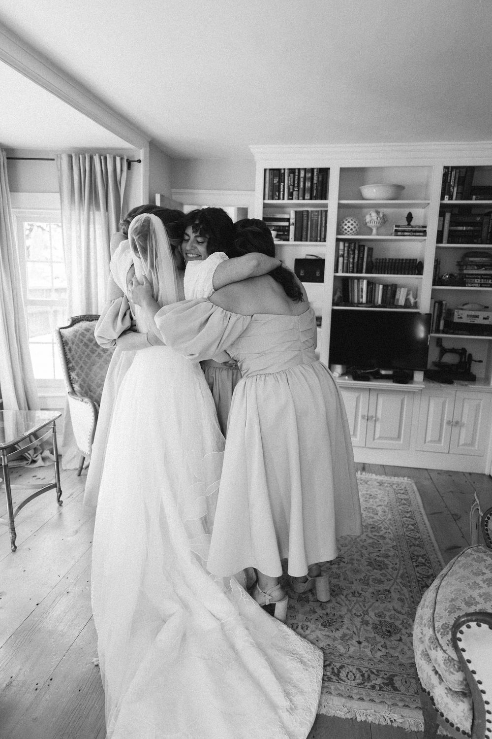 Black and white photo of bride sharing emotional group hug with bridesmaids in cozy room interior.