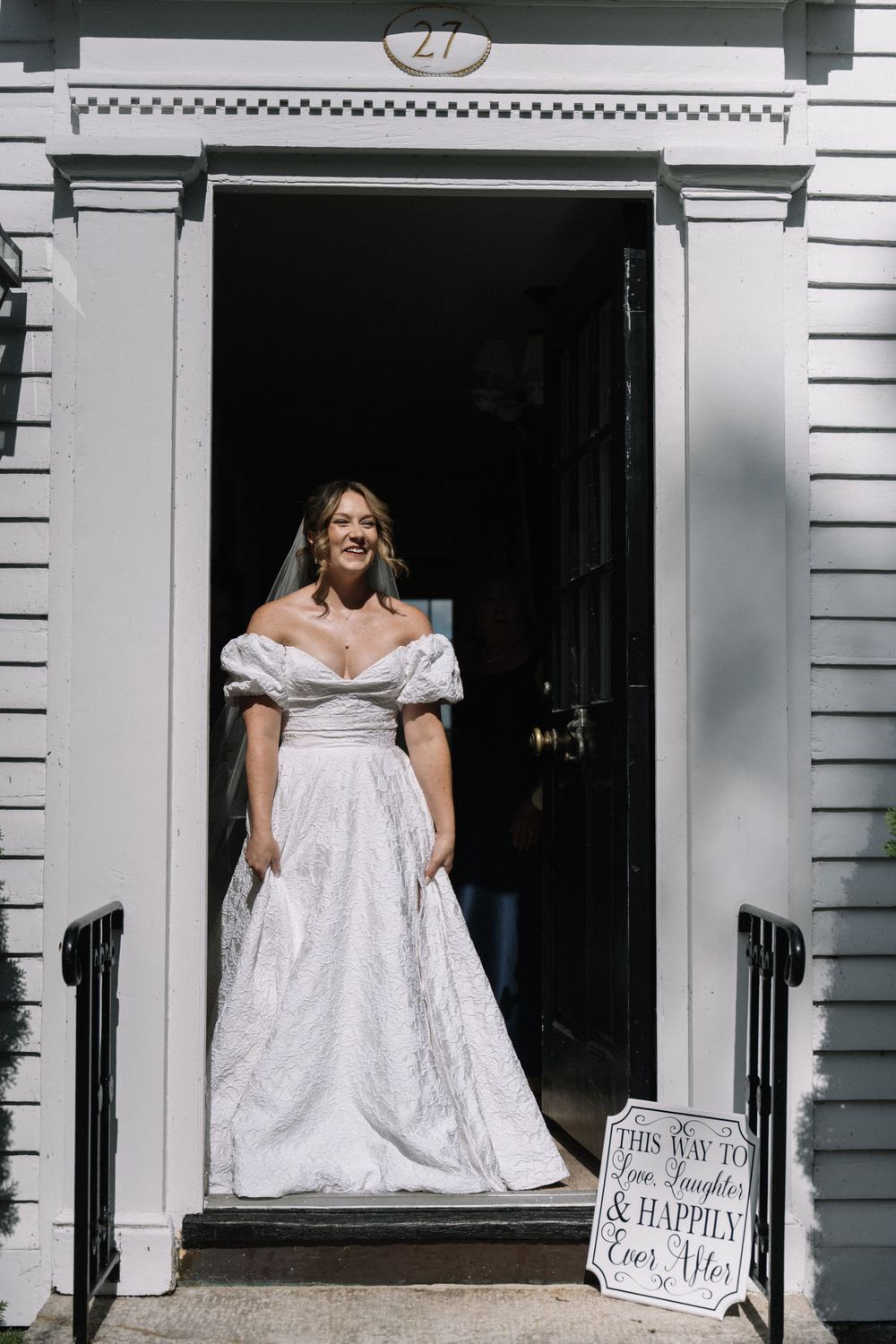 Smiling bride in an off-shoulder white ball gown standing in a doorway with a happily ever after chalkboard sign.