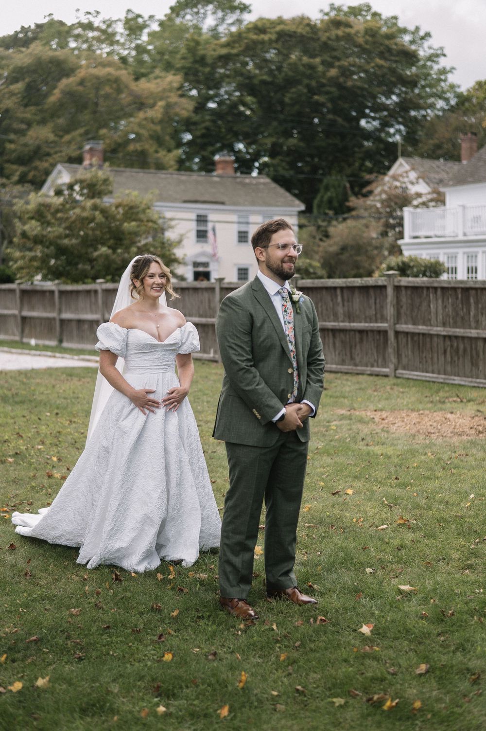 Bride in off-shoulder lace ball gown and groom in green suit stand together on lawn during first look.