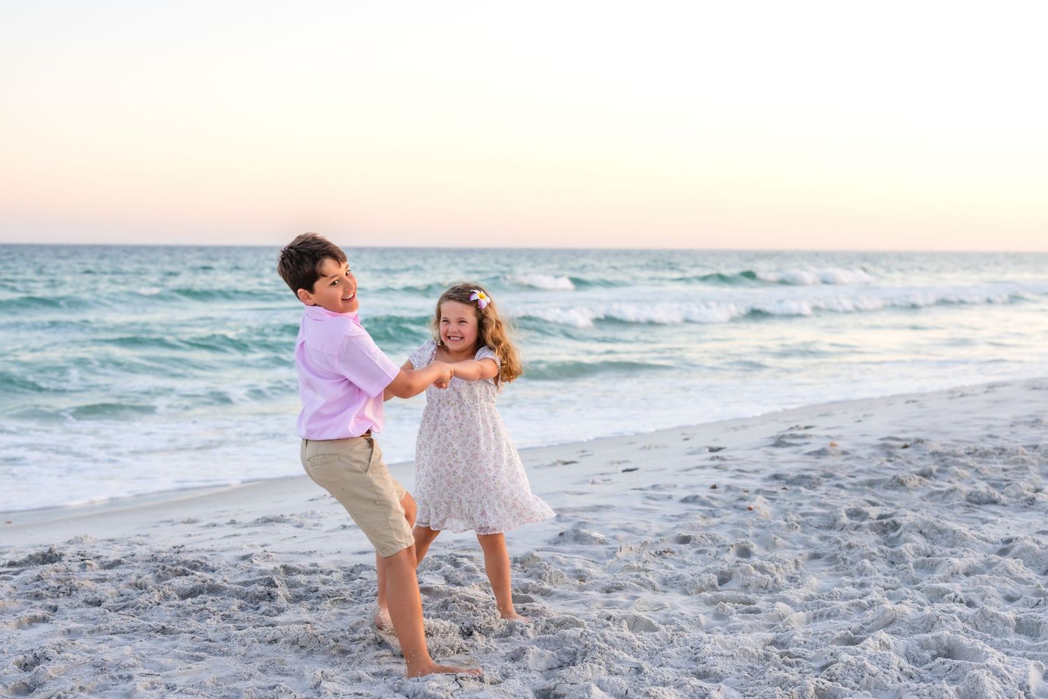 Two children dancing joyfully on a sandy beach at sunset with turquoise ocean waves in the background.