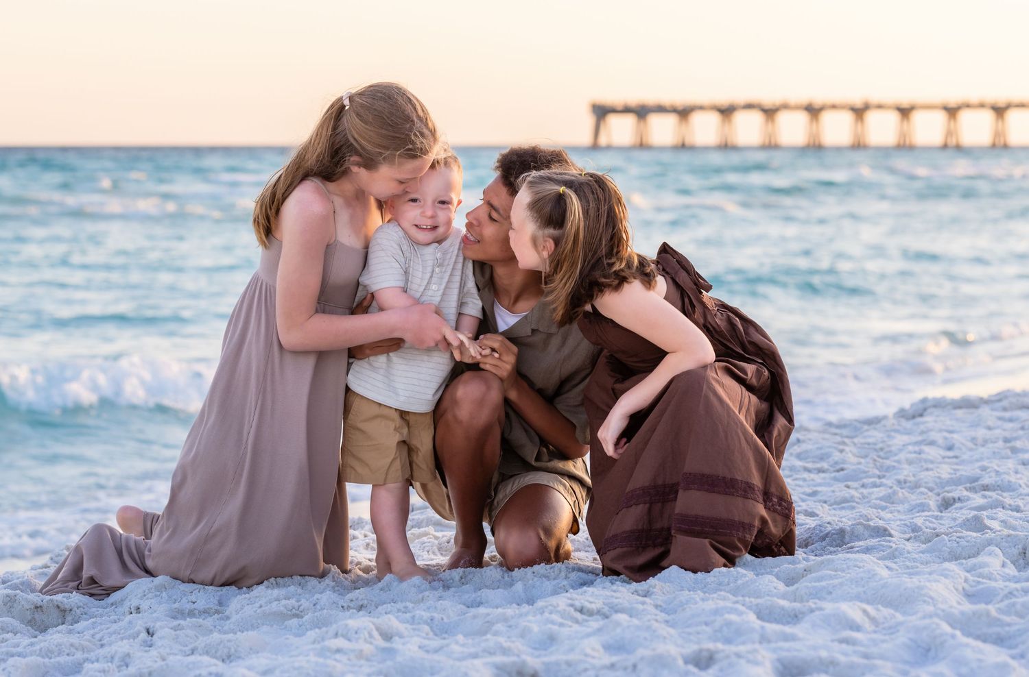 Family posing together on a white sandy beach at sunset with calm turquoise ocean waves in the background.