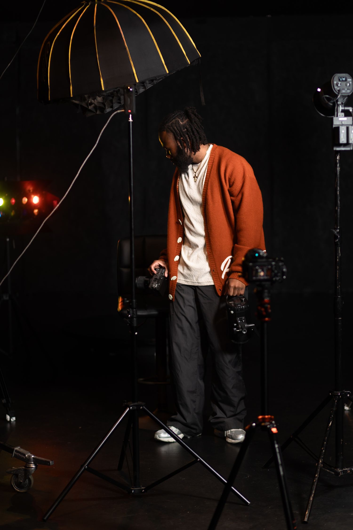A photographer holds two cameras under studio lighting with a softbox overhead in a dark studio setting.