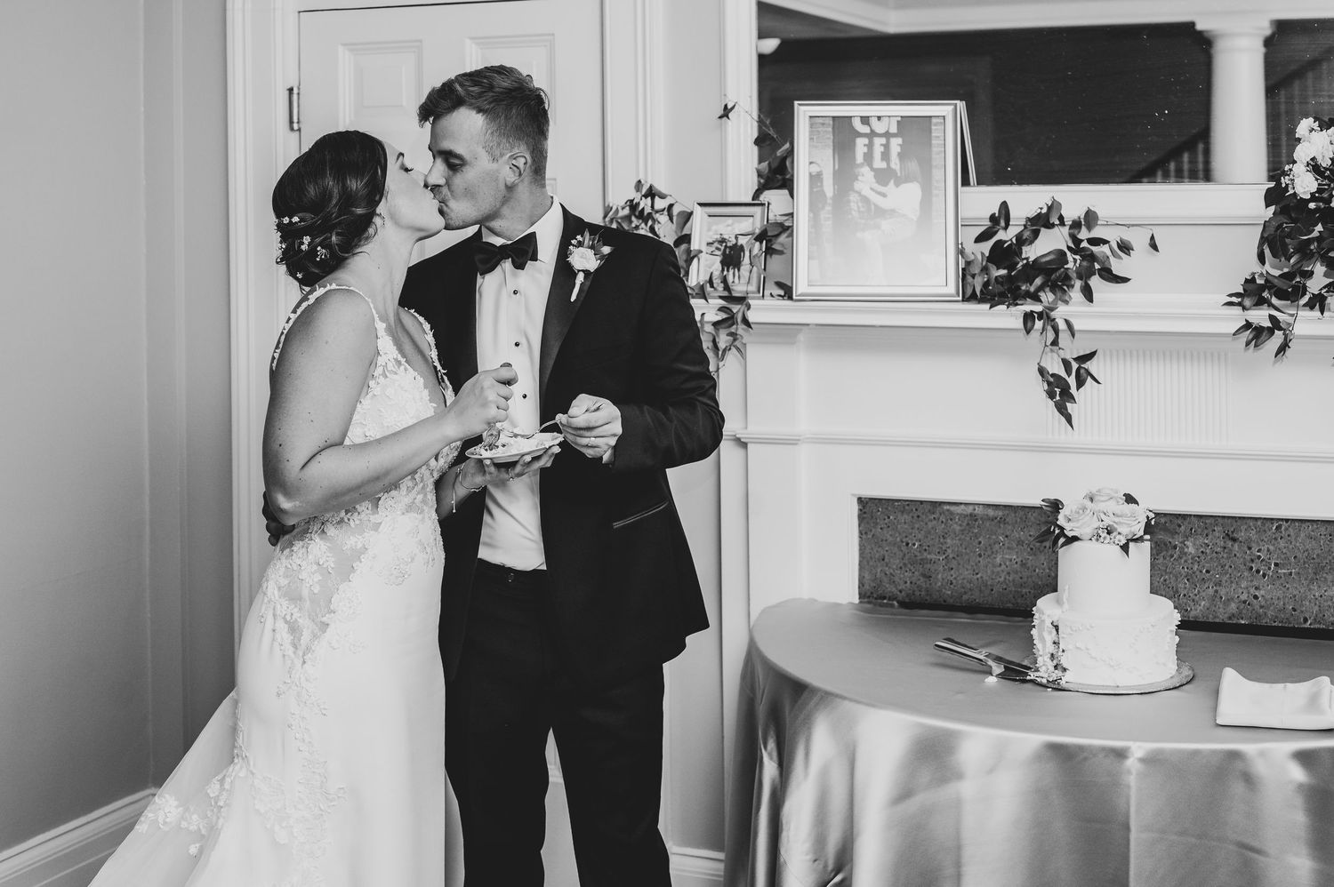 Black and white photo of bride and groom sharing a kiss while cutting their wedding cake at reception.