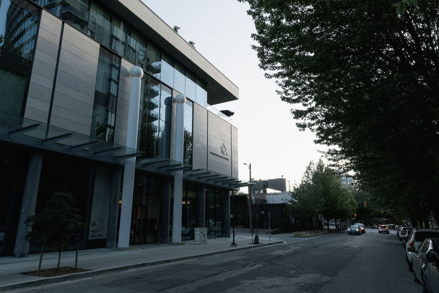 Modern glass-facade event venue building flanked by tall trees along a quiet urban street at dusk.