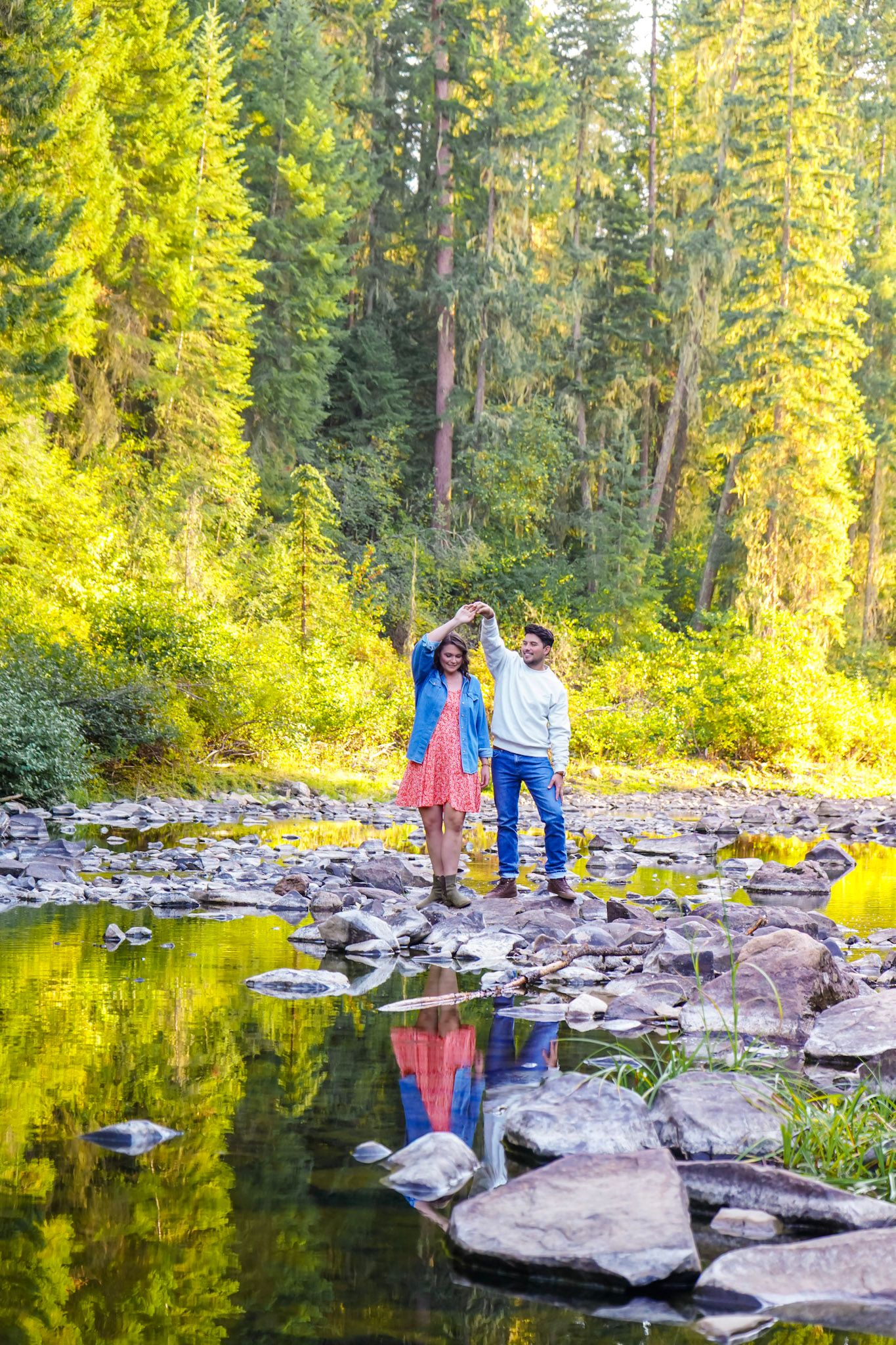 Couple dancing on rocks by a reflective forest stream surrounded by tall evergreen trees in golden sunlight.