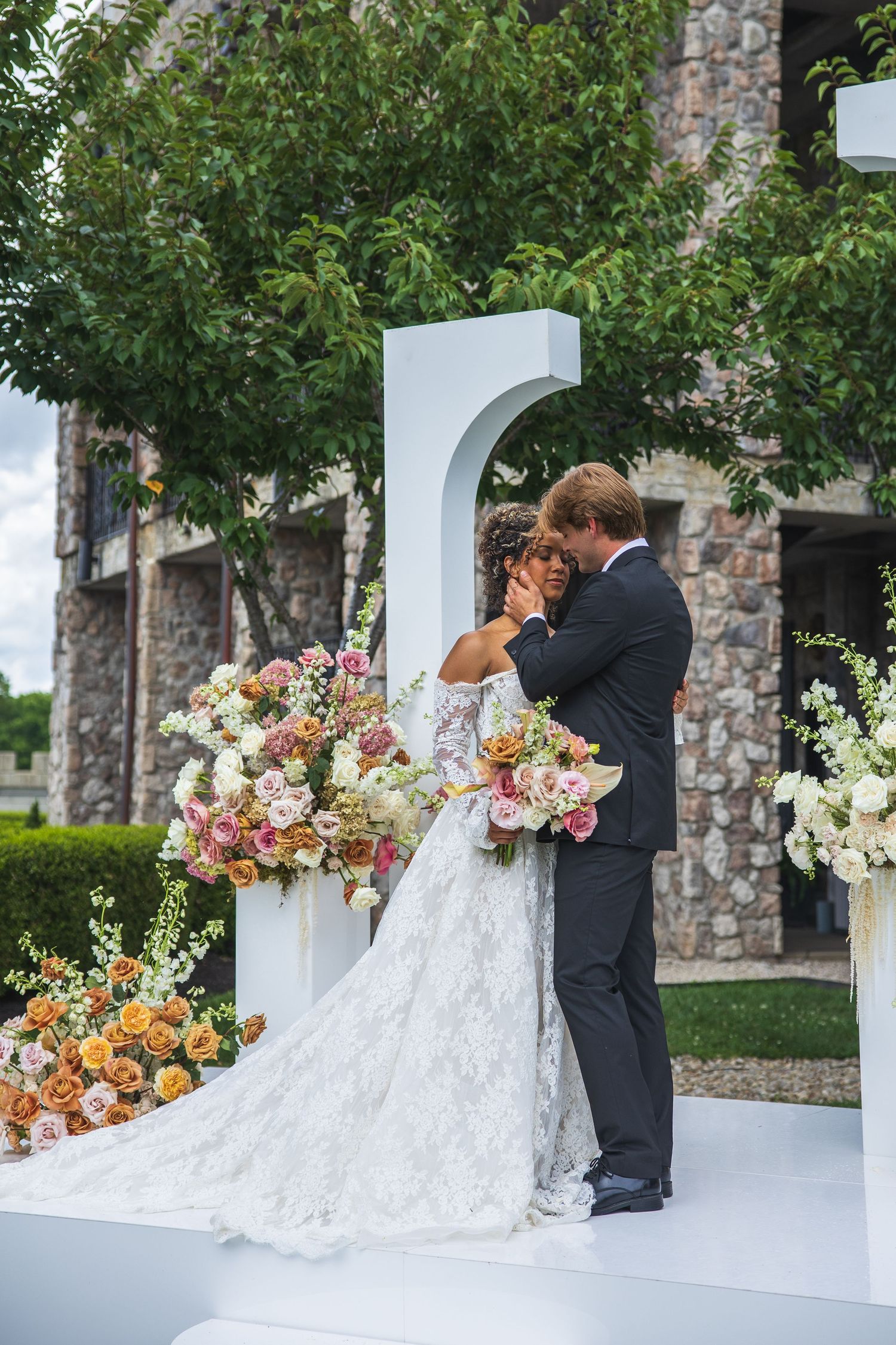 A bride and groom share a romantic embrace at an outdoor wedding ceremony surrounded by lush floral arrangements.