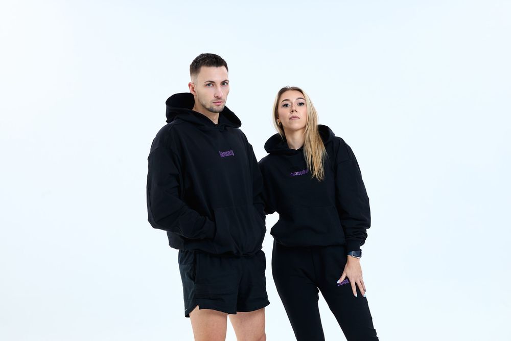 Man and woman wearing matching black oversized hoodies with purple logo print, posing against white background.