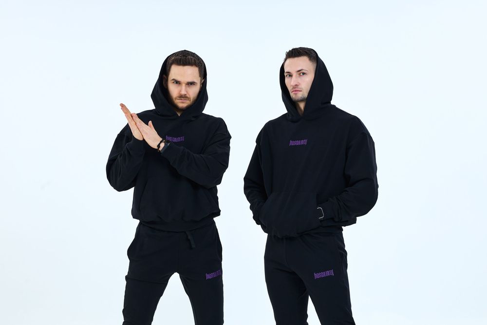 Two men in matching black hoodies and joggers pose against a white background, one crossing arms.