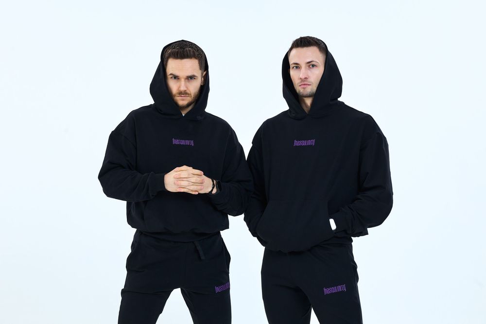 Two men wearing matching black hoodies and sweatpants with purple logo print, posing against a white background.