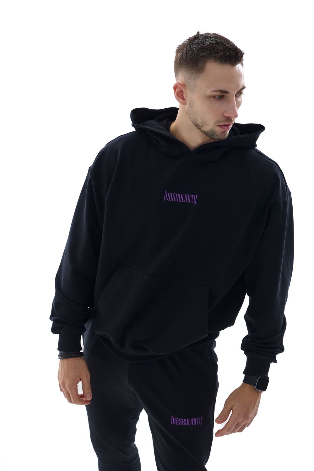 Man wearing black oversized hoodie and matching joggers with purple logo branding on white background.