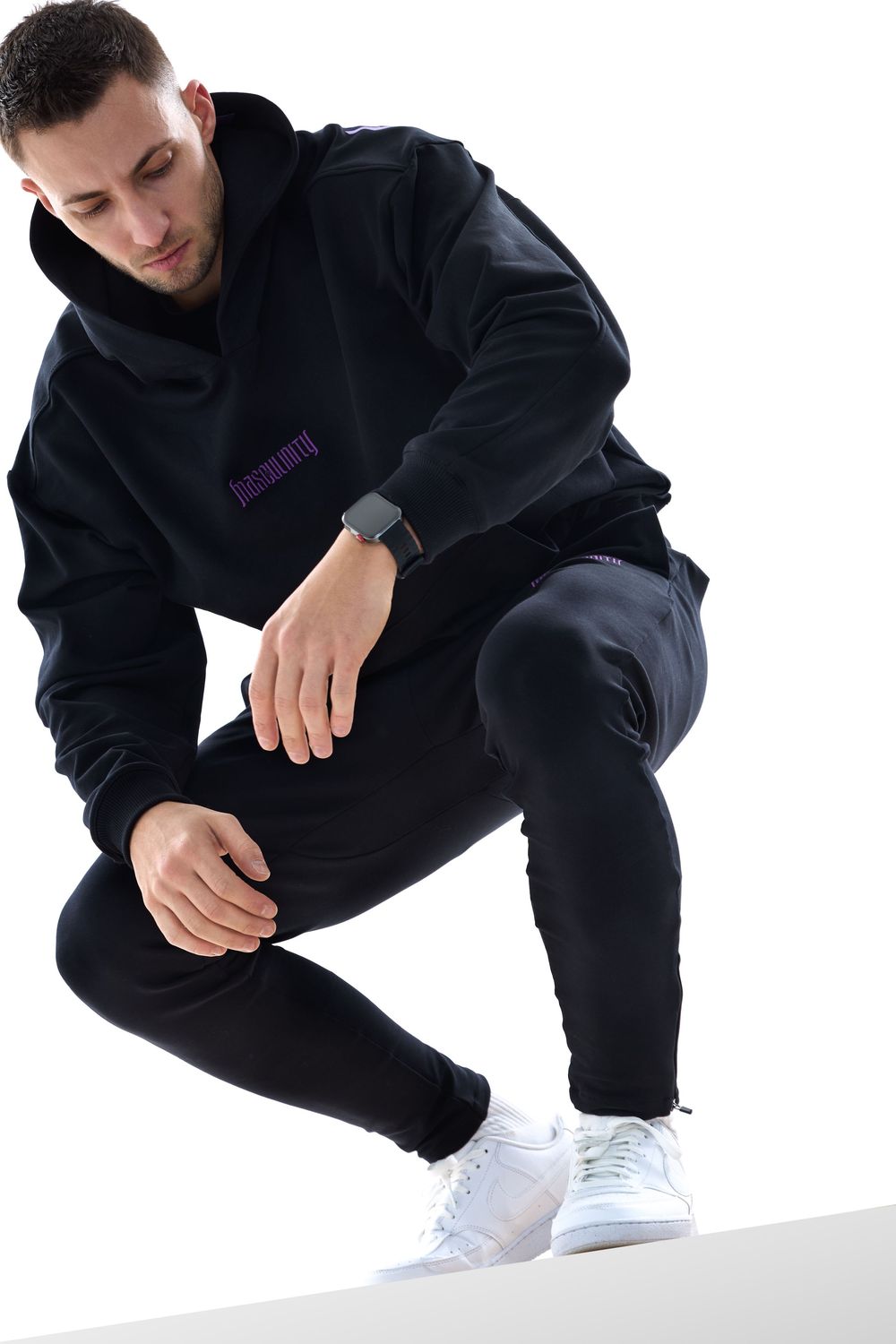 Man in black hoodie and joggers crouching down, wearing white sneakers and a smartwatch on white background.