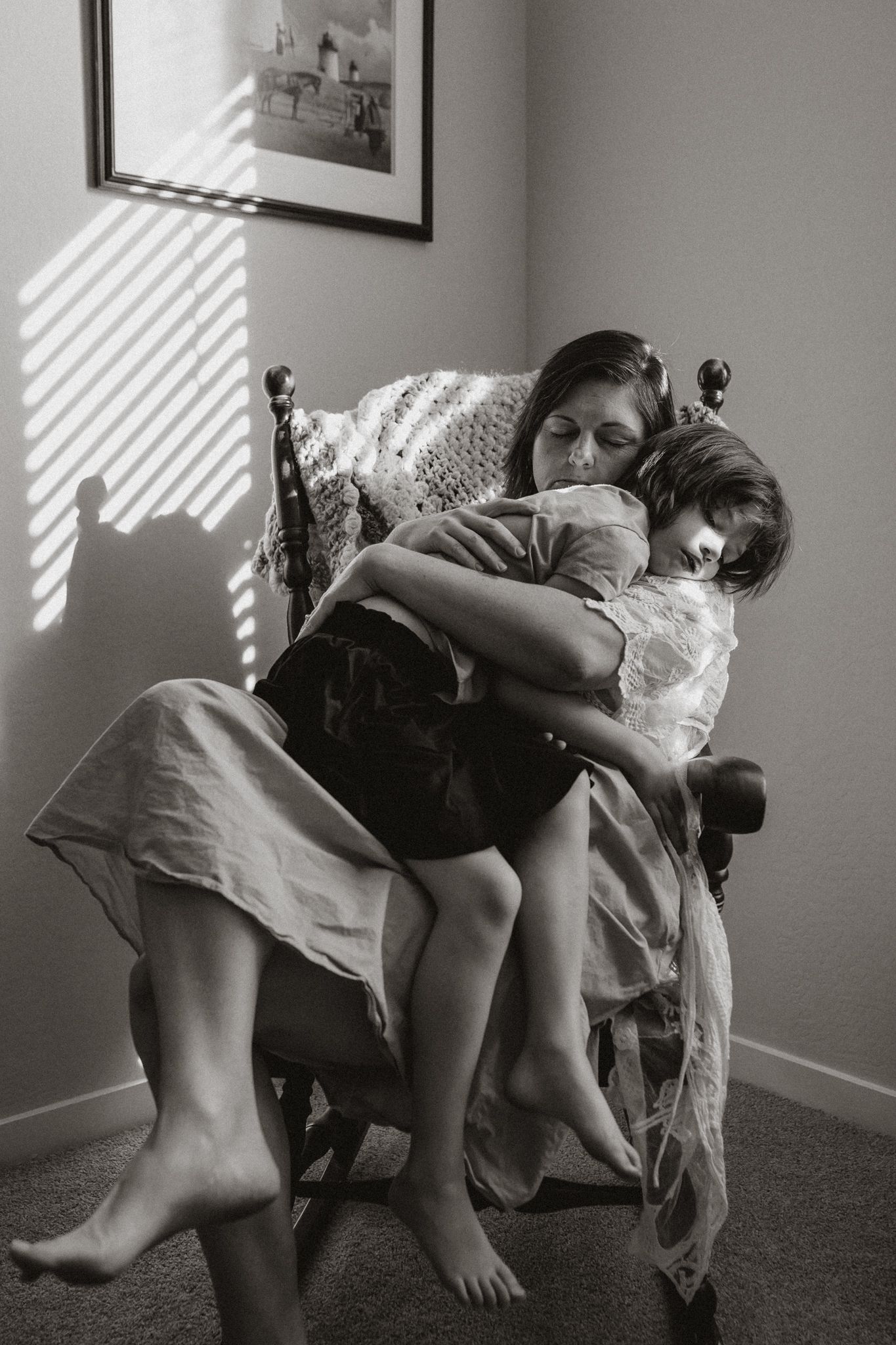 Black and white photo of a mother cradling her child in a wooden rocking chair by sunlit window. Whitney Popp, Phoenix Arizona photographer
