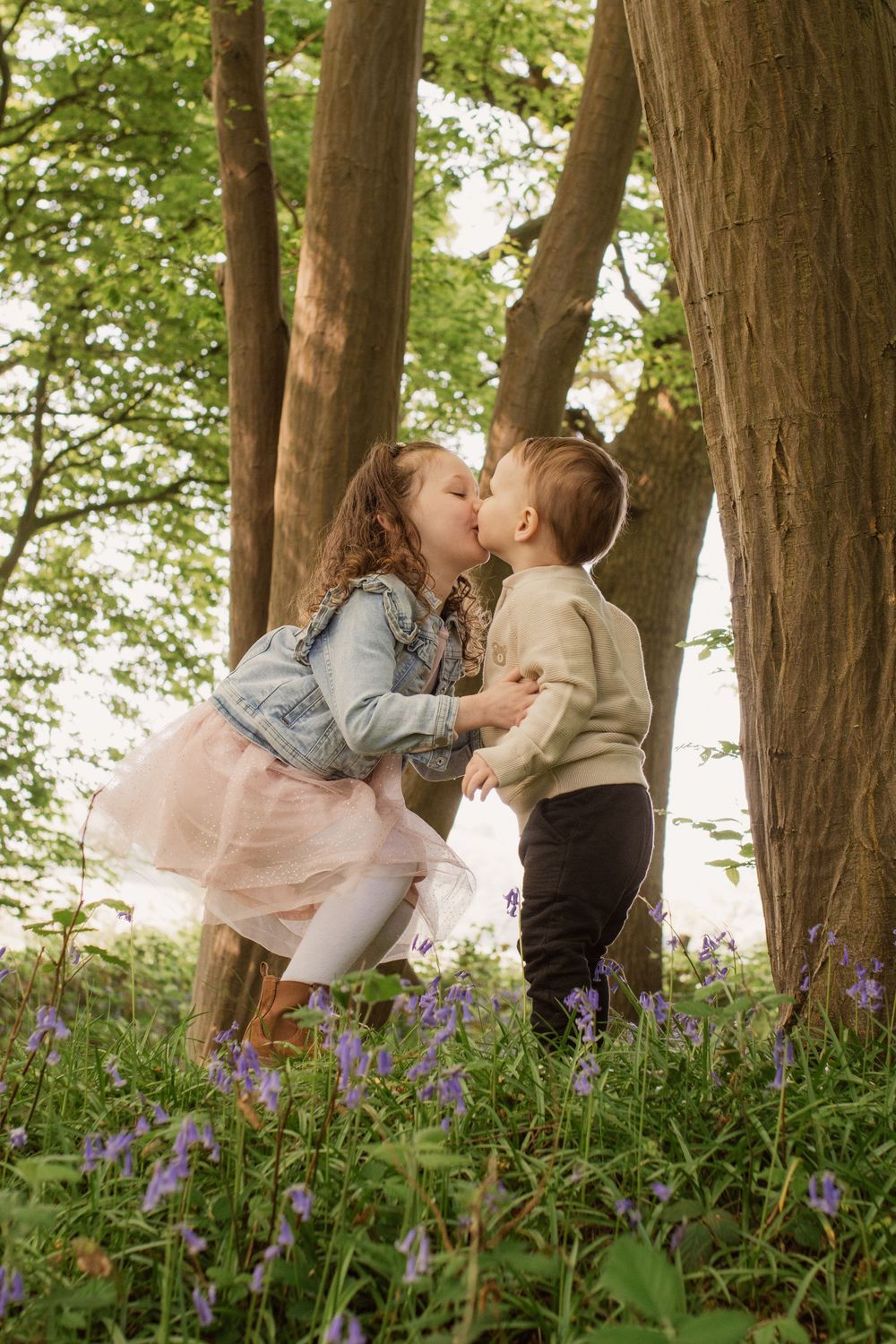 Two young children sharing a sweet kiss among bluebells in a woodland setting.