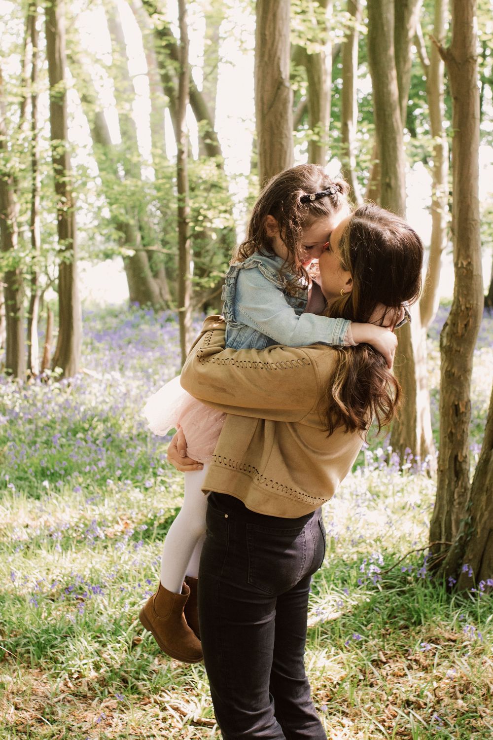 A mother holds her young daughter in a sunlit bluebell woodland, sharing a tender nose-to-nose moment.