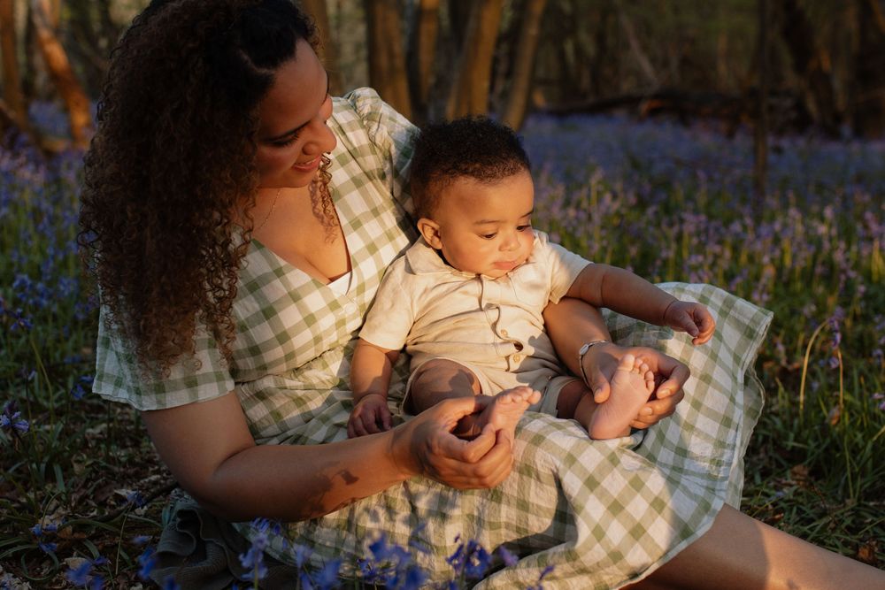 Mother sitting in bluebell field holding baby in golden hour light, wearing green gingham dress.