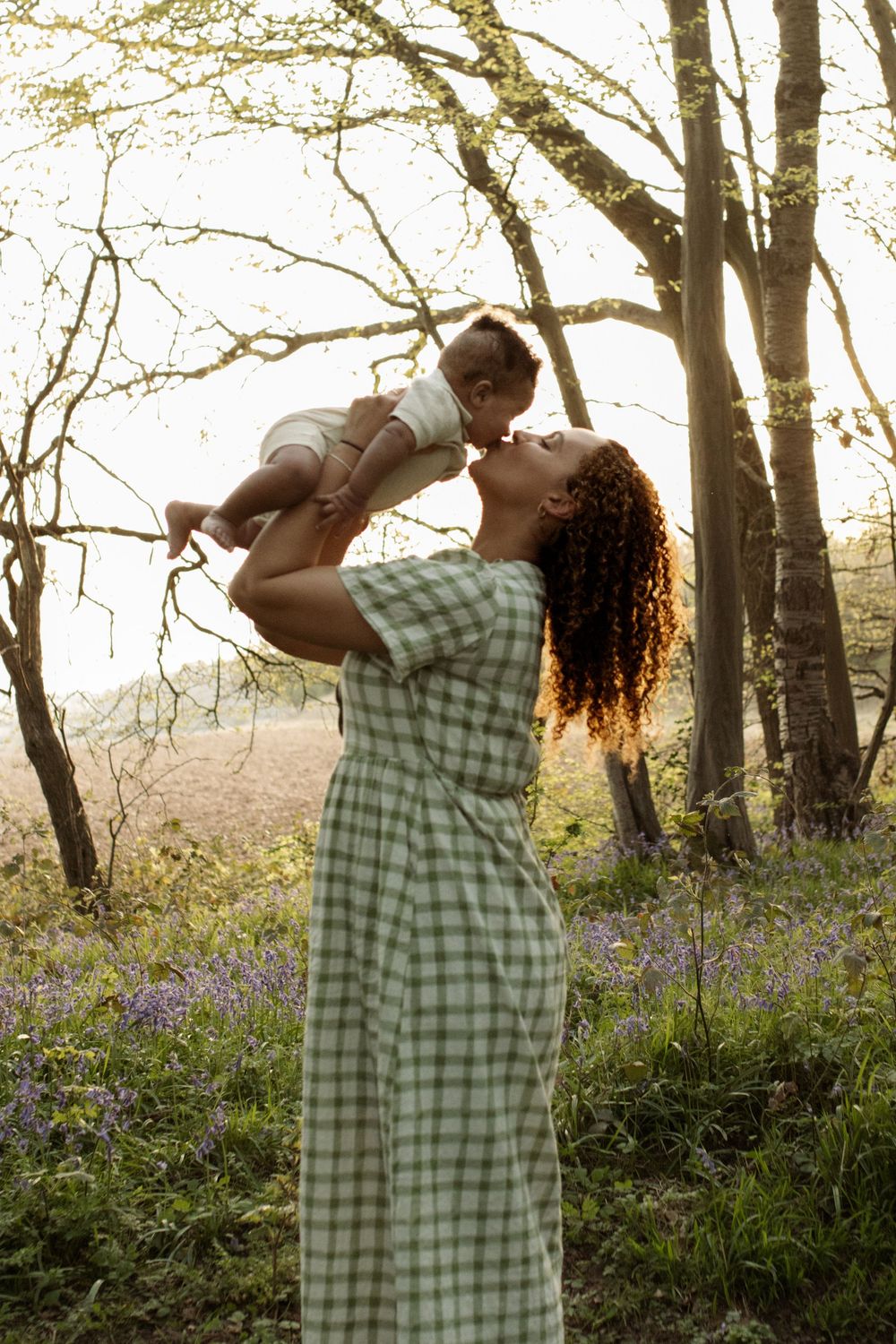 A mother in a green plaid dress lifts her baby up for a kiss in a sunlit forest with bluebells.