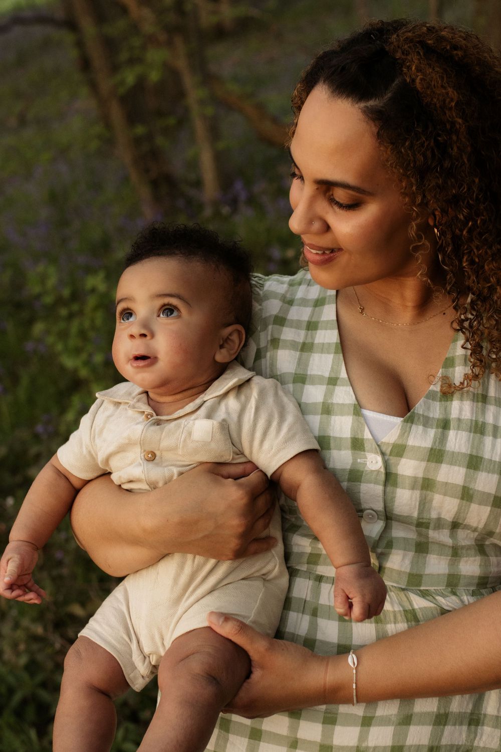 A young mother in a green gingham dress holds her baby boy outdoors among purple wildflowers in a sunlit forest.