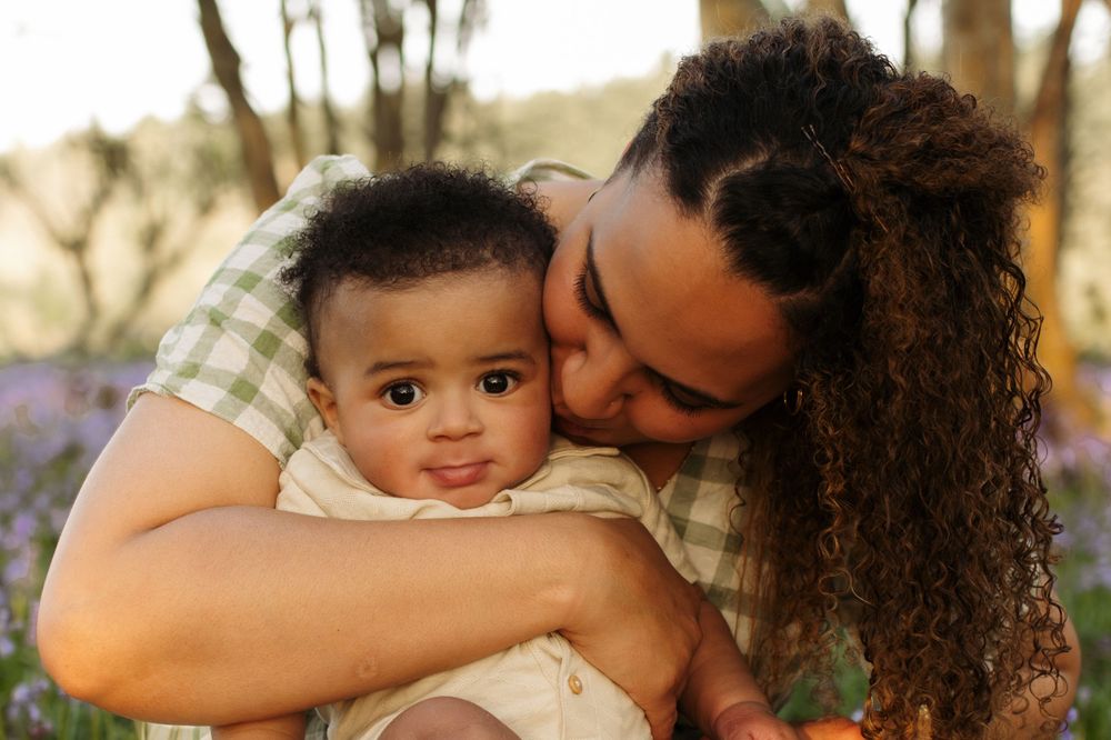 A mother with curly hair tenderly kisses her baby outdoors in a sunlit park with purple flowers.