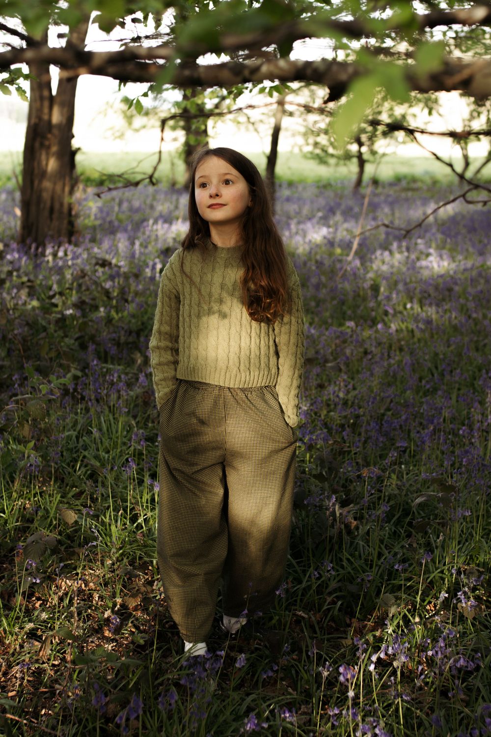 A young girl in olive green knit sweater and trousers stands among purple bluebells in a woodland setting.