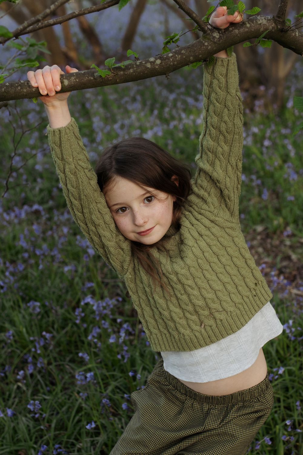 Young girl in green cable-knit sweater hanging from tree branch in field of purple wildflowers.
