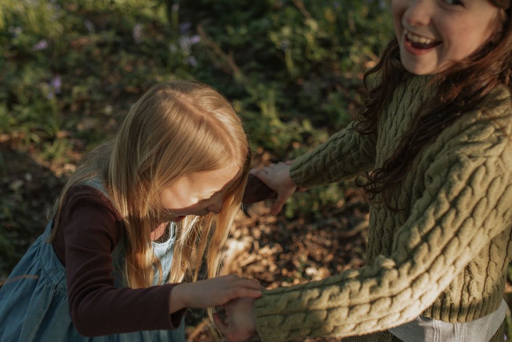 A young girl and a smiling woman in a green knit sweater play together outdoors among lush greenery.