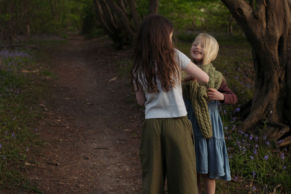 Two young girls hugging on a forest path surrounded by lush green trees in soft natural light.