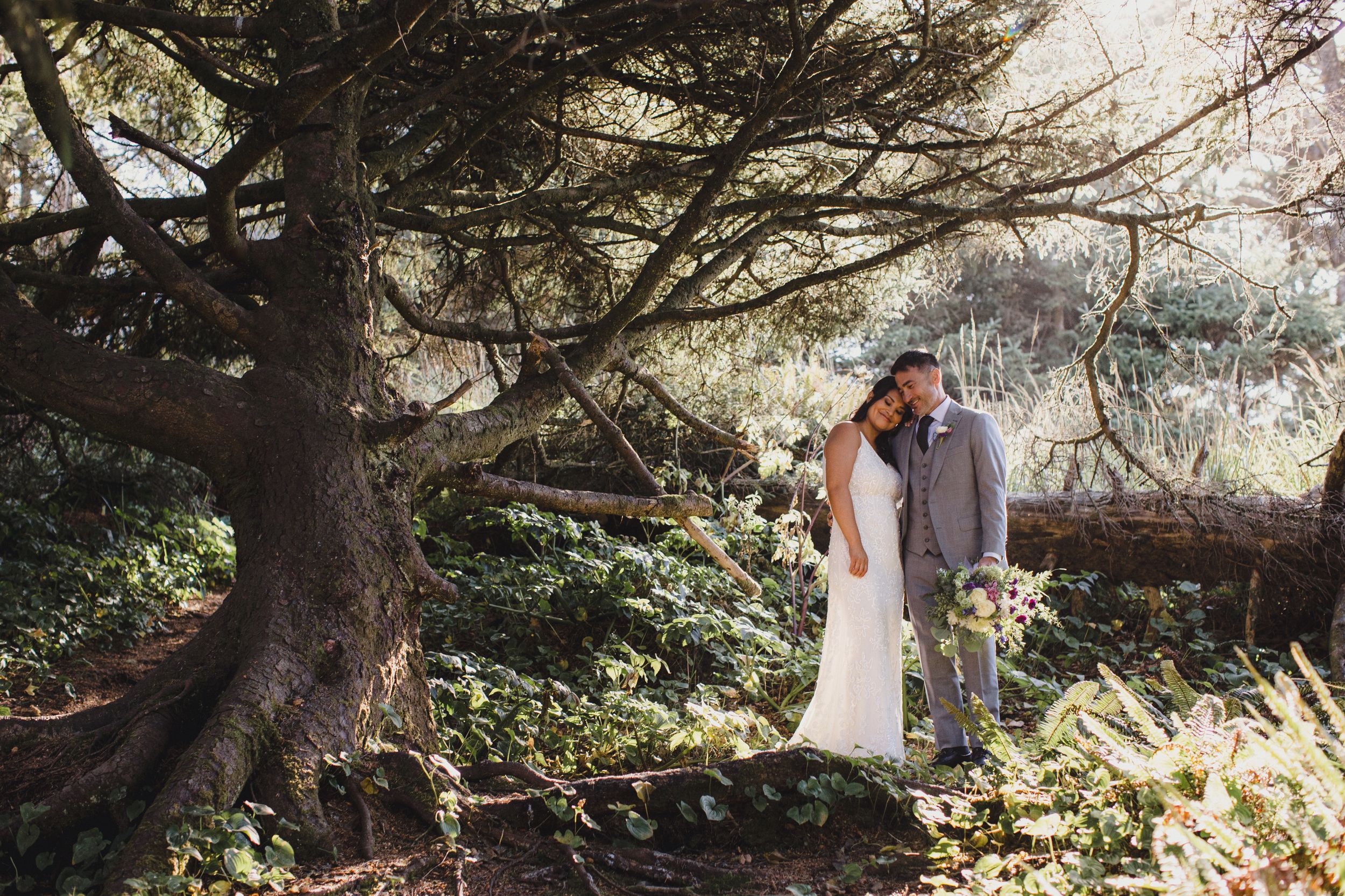 Bride and groom embracing under a large tree in a lush forest setting during their outdoor wedding ceremony.