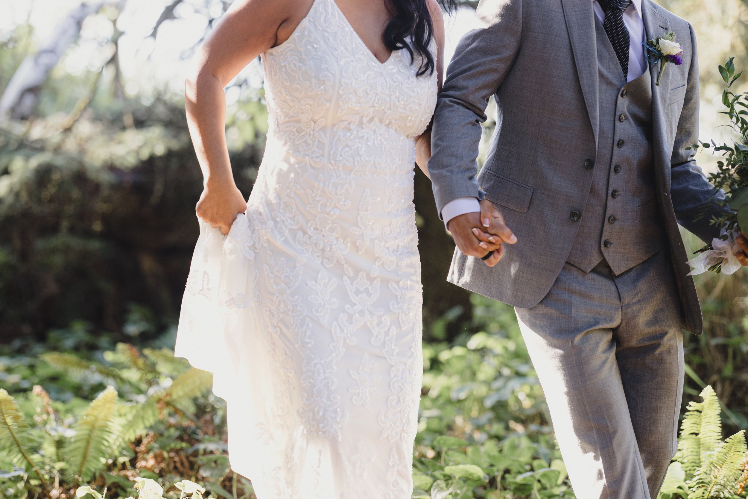 Bride in white lace gown and groom in gray suit holding hands outdoors among lush green ferns.