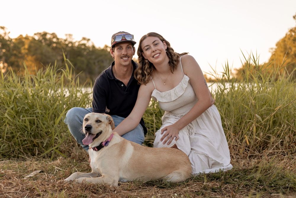 Couple smiling with golden Labrador dog outdoors in golden hour sunlight near tall grass field.