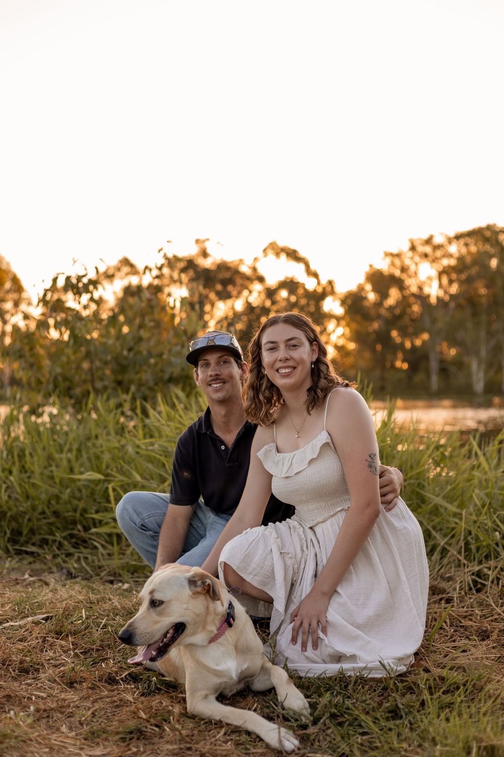 Couple sitting in golden hour sunlight with their yellow Labrador in a lush green field near a lake.